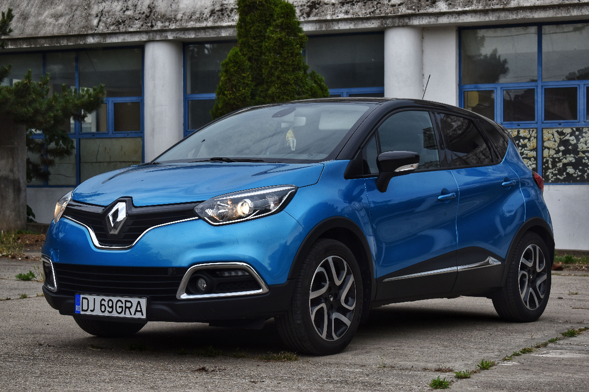 Mavi renkte Renault markalı otomobil, önünde ve yanlarında siyah detaylar, arka planda mavi çerçeveli pencereler ve beton bina, araç ön sol açıdan görünür halde park halinde.