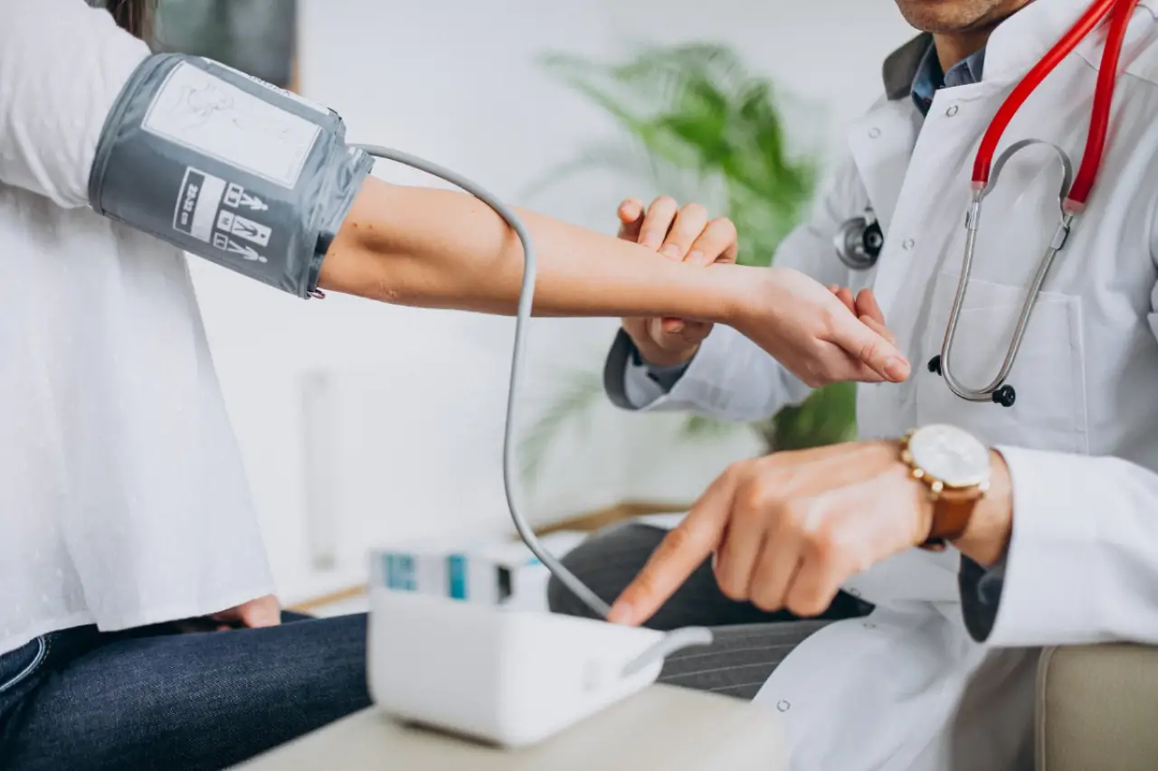 Beyaz önlük giymiş bir doktor, hastanın koluna tansiyon aleti manşetini takarken diğer eliyle hastanın nabzını kontrol ediyor.