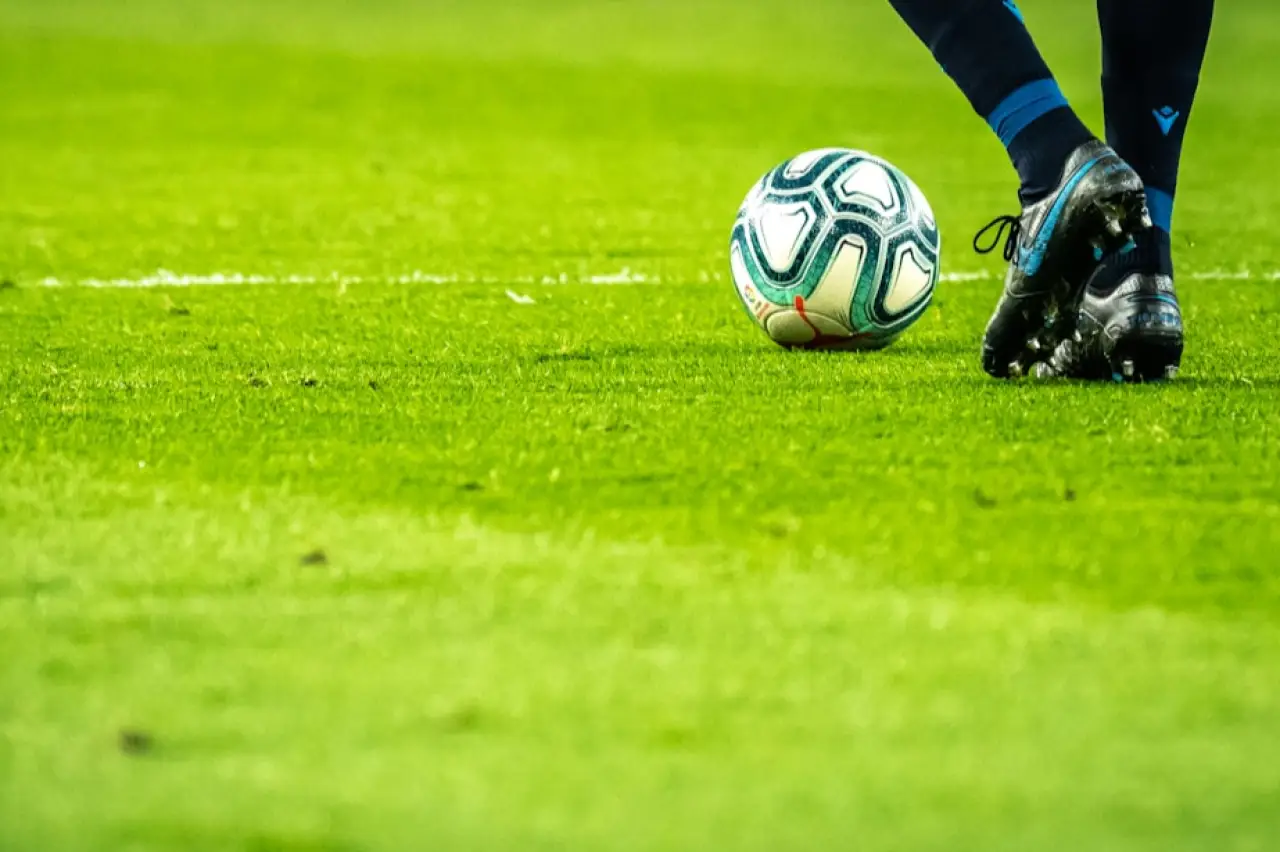 Yeşil çim sahada, siyah ve mavi futbol ayakkabısı giymiş bir kişinin topa vurmaya hazırlandığı futbol topu yakın çekim görüntüsü.