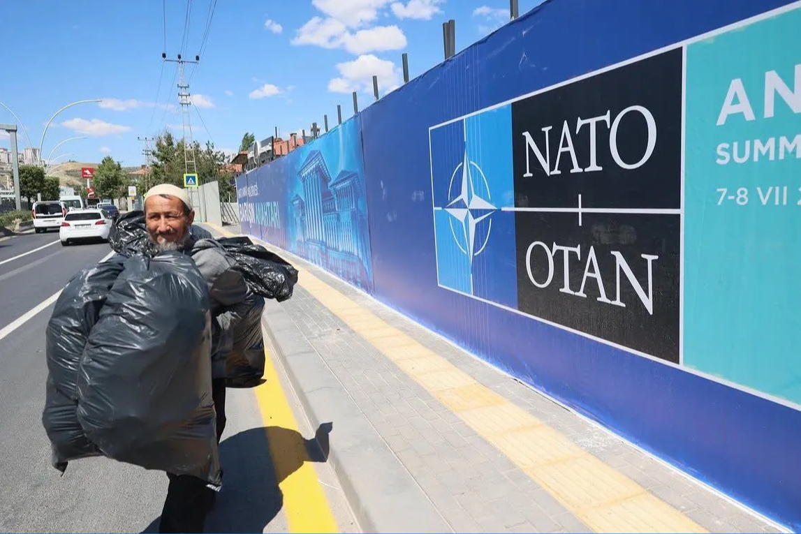 Bir erkek, iki büyük siyah plastik çöp torbasını omzunda taşıyarak kaldırımla yol arasındaki alanda yürüyor, arkasında NATO yazılı büyük pankart bulunuyor, şehir içi araçların geçtiği cadde ve mavi gökyüzü görünüyor.