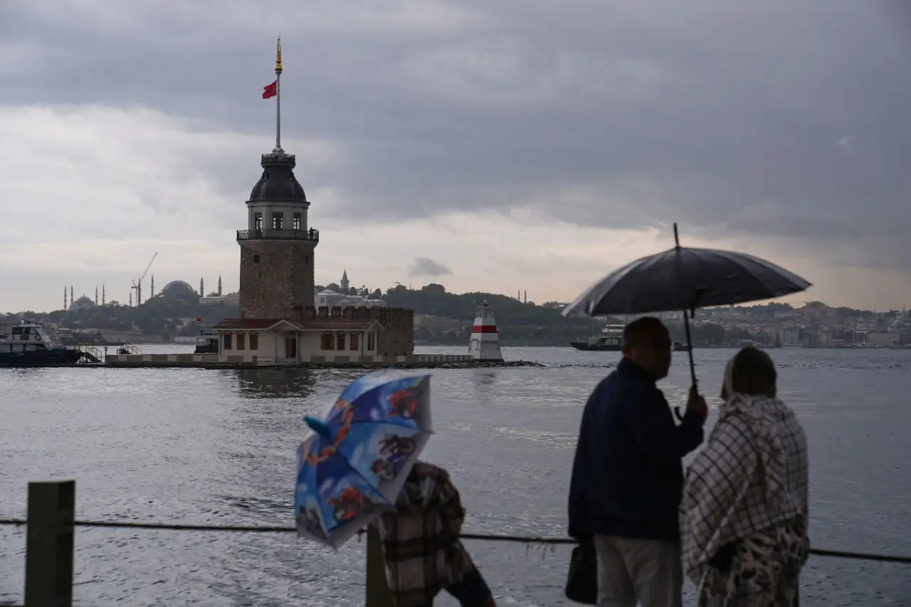 İki kişi, kıyıda bir gözetleme kulesinin önünde dururken yağmurluk ve şemsiye kullanıyor. Arkada, İstanbul silueti ve çeşitli cami minareleri görünür. Hava kapalı ve suyun üzerinde hafif dalgalar var