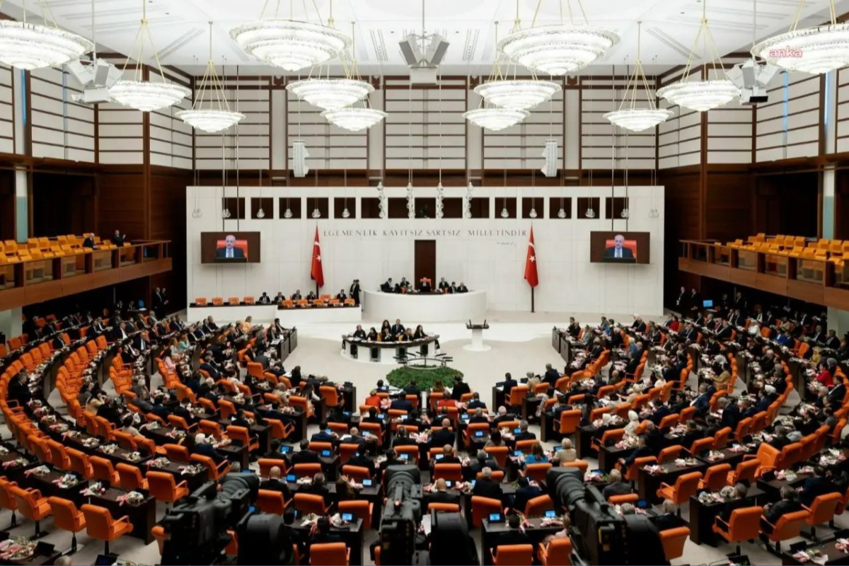 Türkiye Büyük Millet Meclisi'nde gerçekleştirilen bir oturum, çeşitli siyasi partilerden milletvekillerinin tartışmalara katıldığı bir ortamda görünmektedir. Salonda turuncu sandalyeler, ekranlar ve Türk bayrakları belirgin şekilde yer almakta. Oturum başkanlığı ise ön planda yer alan bir masadan yönetilmektedir. Meclis, TBMM