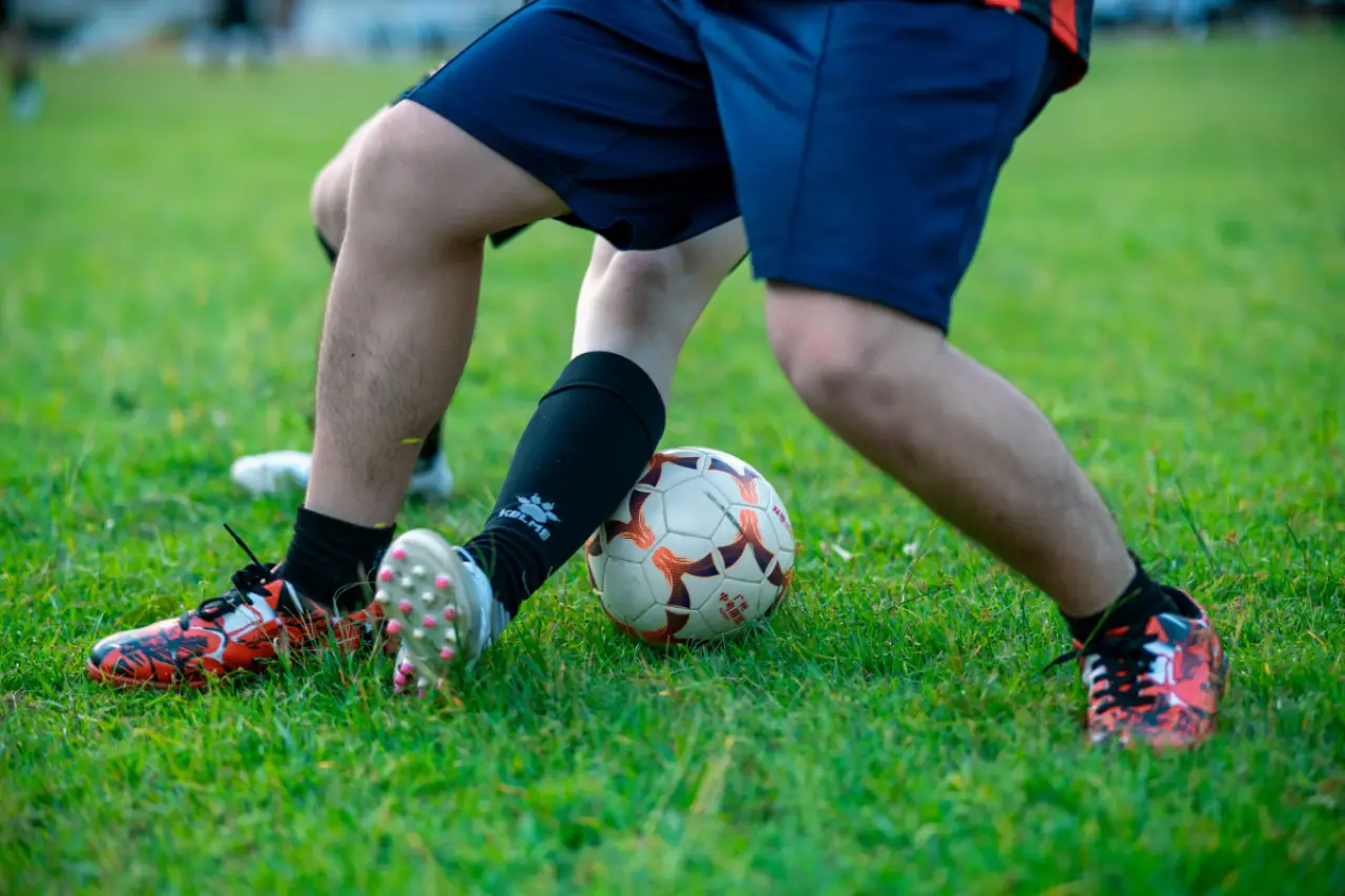 İki futbolcunun, çim alanda bir futbol topu etrafında mücadele ettiği bir yakın plan. Bir oyuncunun kırmızı ve siyah futbol ayakkabıları, diğerinin ise siyah çorap ve beyaz ayakkabıları dikkat çekiyor. Top, oyuncuların ayakları arasında konumlanmış.