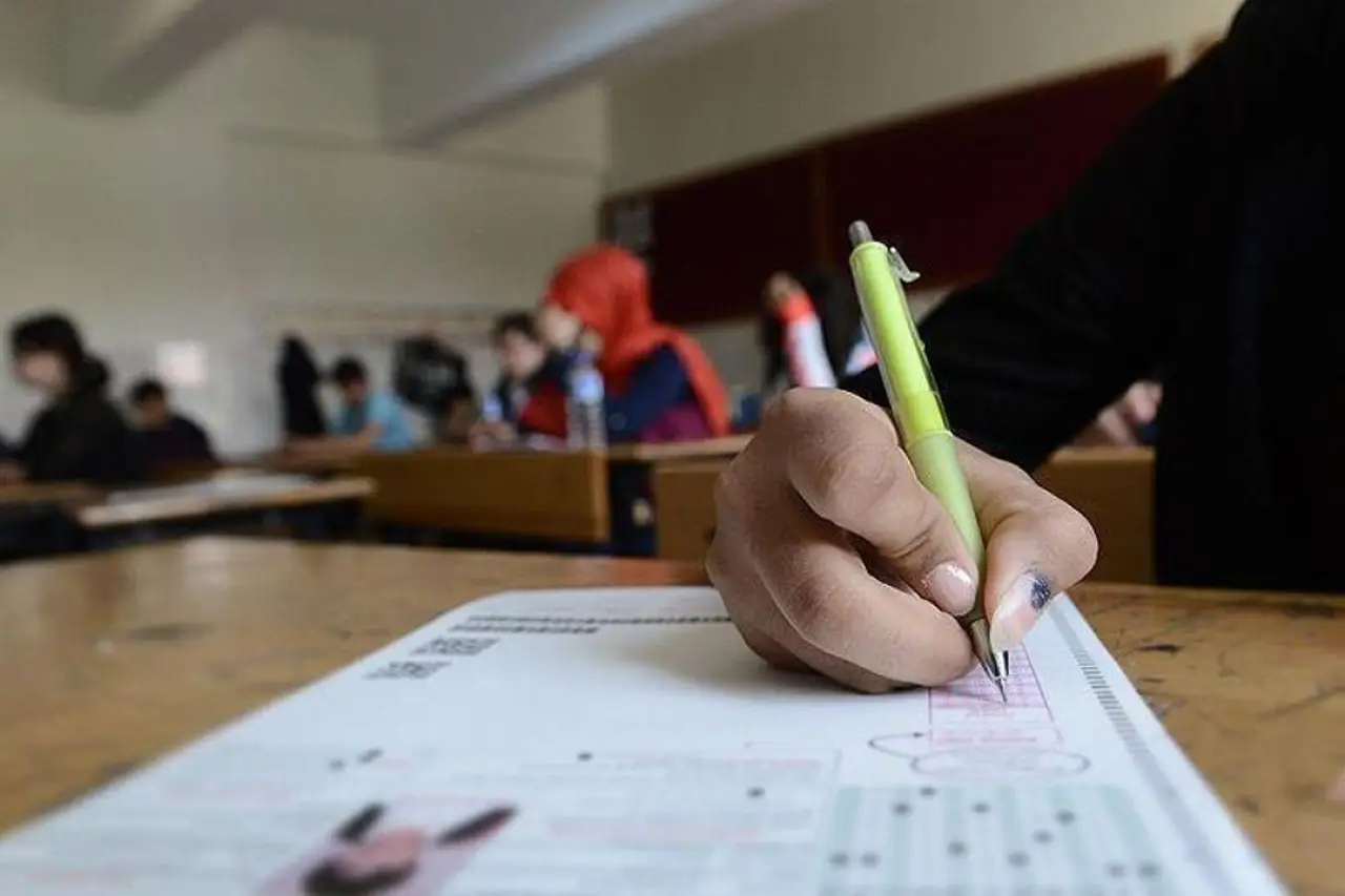 Bir öğrencinin, yeşil bir kalemle sınav kağıdına yazı yazdığı bir sınıf ortamı. Arkada diğer öğrenciler sınavda. Sınıf, hafif loş bir ışıkta, ahşap sıralar ve beyaz duvarlarla donatılmış.
