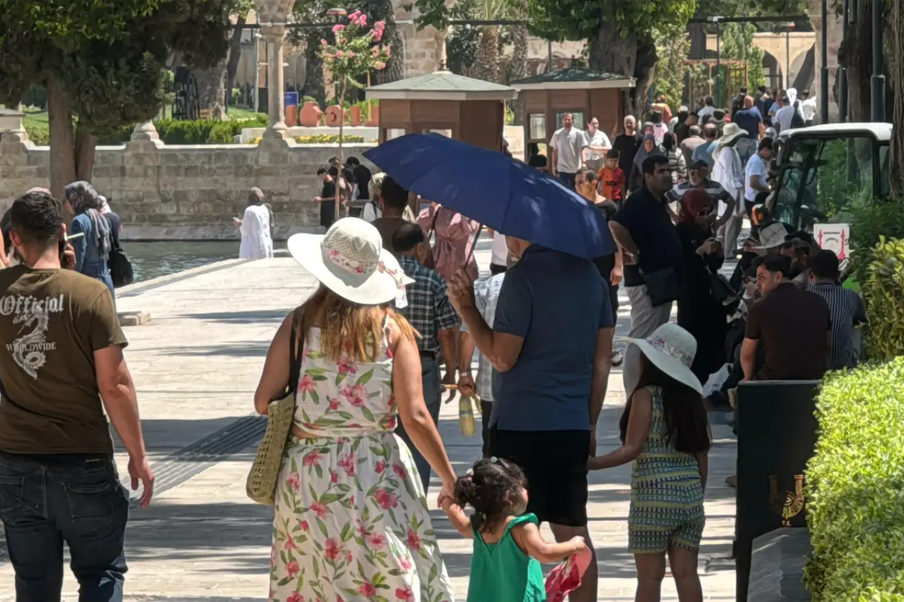 Kalabalık bir sokakta yürüyen çeşitli insanlar görünmektedir. İki kadın, bir kız çocuğuyla birlikte yürüyor; biri çiçekli elbise, diğeri yeşil elbise giymekte. Arkada, bir grup kişi iğne iplik yürüyüş yapıyor. Güneşten korunmak için şemsiyeler kullanılıyor.