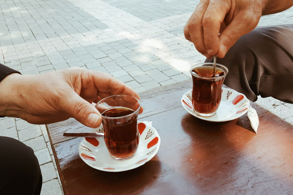 İki el, masada duran iki küçük çay bardağında çay karıştırırken görünmektedir. Bardağının yanında beyaz ve kırmızı desenli tabaklar, masanın üzerinde ise taş döşeme görülebilir.