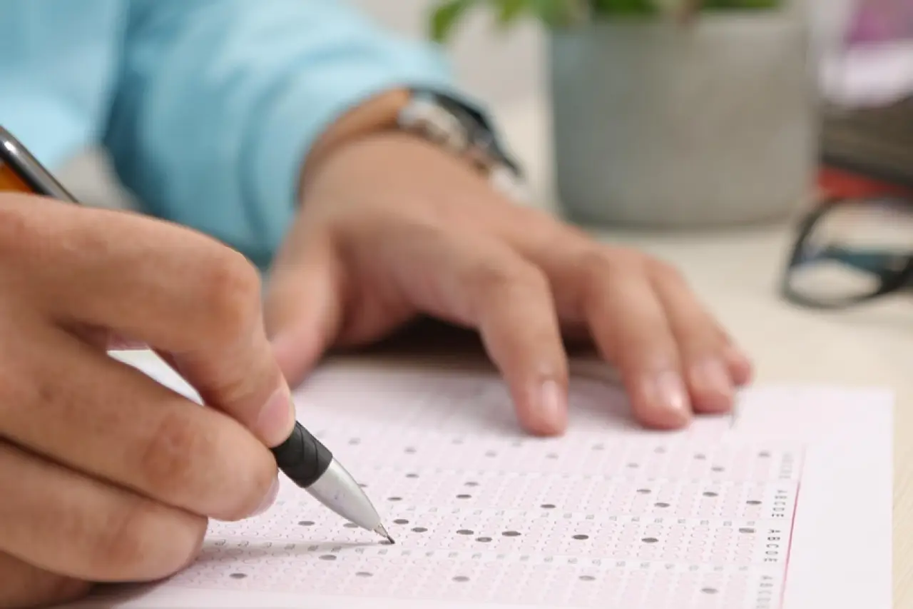 Bir kişi, bir masada test formunu doldururken bir kalemle işaretlemeler yapıyor. Masanın arkasında bir saksıda yeşil bir bitki ve yanında gözlük ile hesap makinesi bulunuyor.
