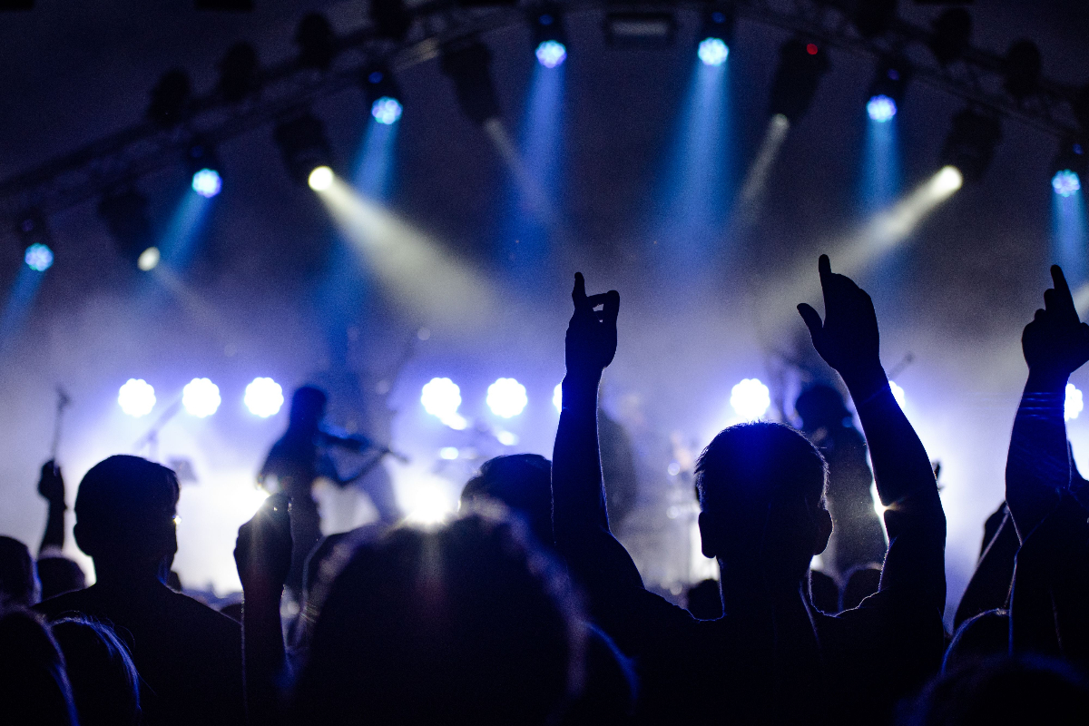 Kalabalık bir konser alanında, izleyicilerin elleri havada, müzik eşliğinde coşkuyla dans ettiği bir an. Arka planda sahnede enstrüman çalan sanatçılar ve parlak ışıklar yer almakta.