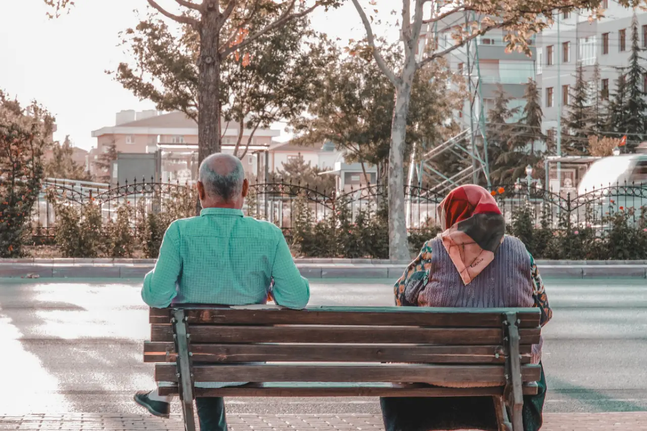 Bir bankta oturan erkek ve kadın, sokakta geçen araçları izliyor. Arka planda binalar, ağaçlar ve çiçekler görünüyor. Kadının başında renkli bir eşarp var, erkeğin ise kısa beyaz saçları dikkat çekiyor.