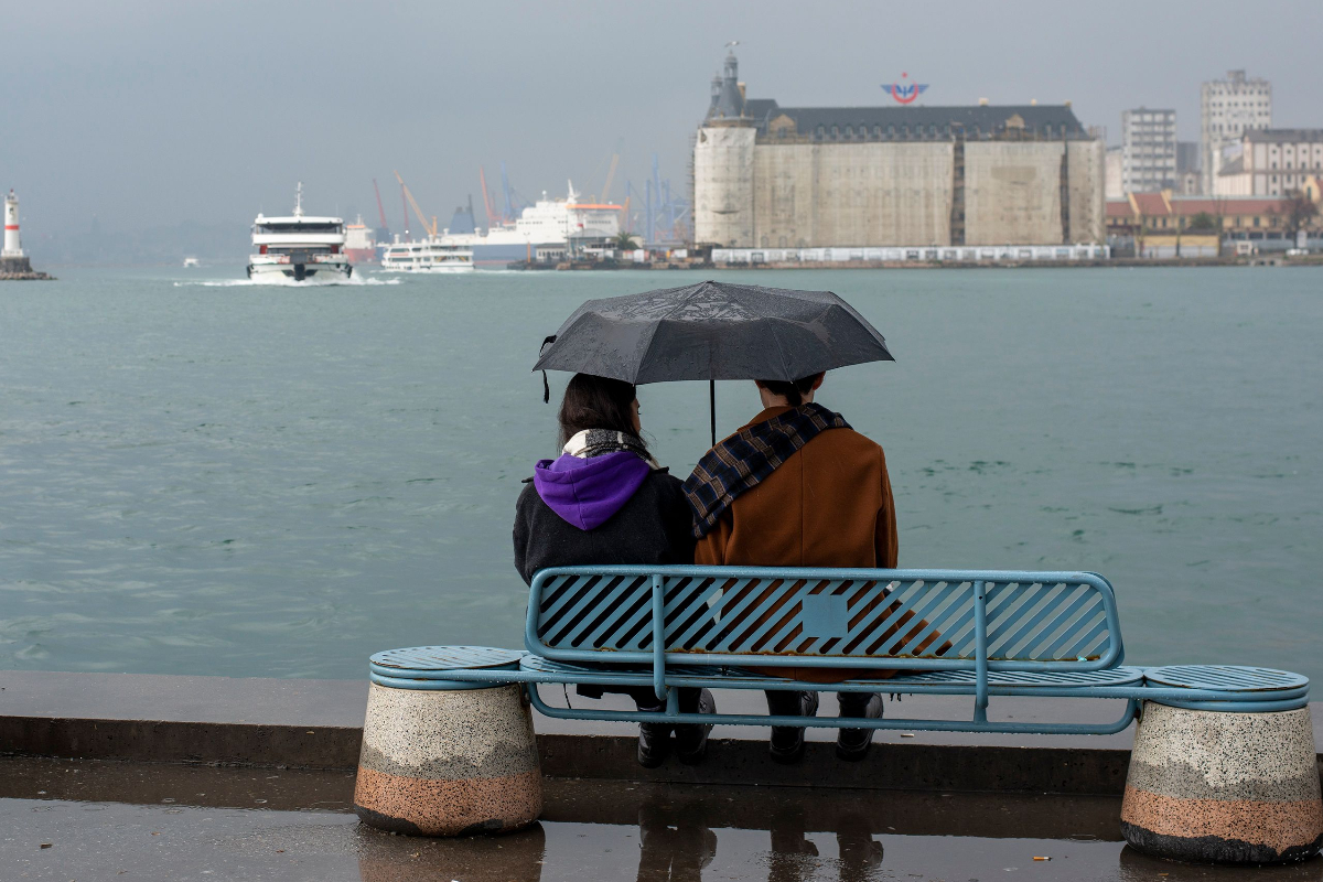 İstanbul'da bir çift elinde şemsiye yağmur altında oturuyor, arkada Haydarpaşa Garı görülüyor
