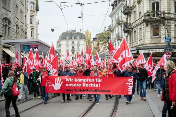 tramvay yolu üzerinde yürüyüş yapan kalabalık işçi grubu ellerinde kırmızı renkte sendika bayrağı tutuyor
