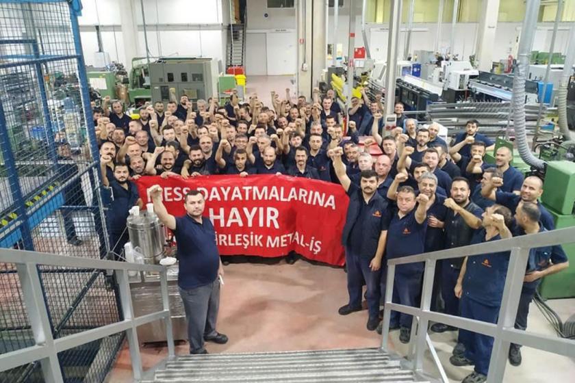 Fabrikada toplanan metal işçilerinin yumrukları havada, ellerinde büyük kırmızı bir pankart var. Pankartta, MESS dayatmalarına hayır, Birleşik Metal-İş yazıyor.