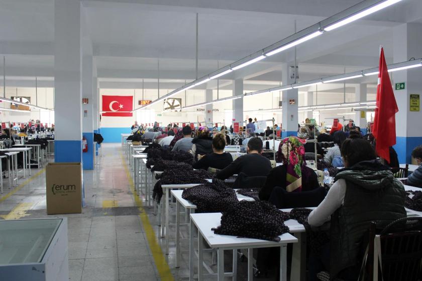 Tekstil atölyesinde işçiler sıra sıra dizilmiş halde çalışırken. İşçilerin çalışıtığı salonun büyük duvarında Türk Bayrağı asılı