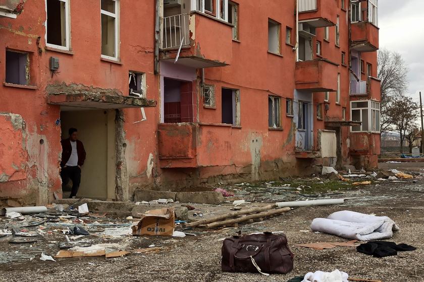 Bazı yerleri yıkılmış bir apartmanın kapısında bekleyen bir adam. Apartmanın önünde yerlere saçılmış koliler, yataklar çantalar...