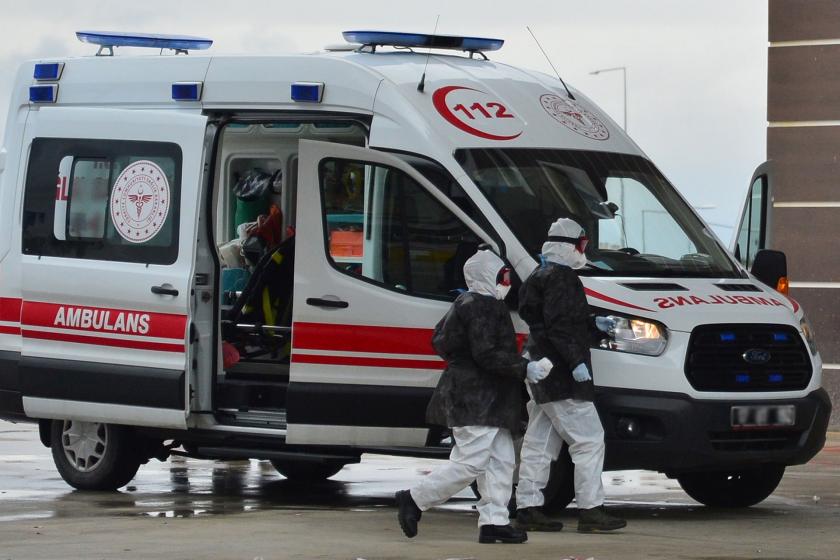 Bir ambulans ve önünden koruyucu elbiseleriyle geçen iki sağlık çalışanı.