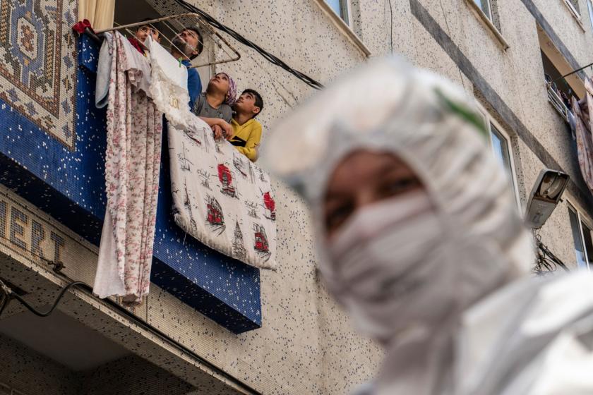 Önde sağda filyasyon ekibinde çalışan bir sağlıkçı, solda balkondan dışarı bakan aile bireyleri