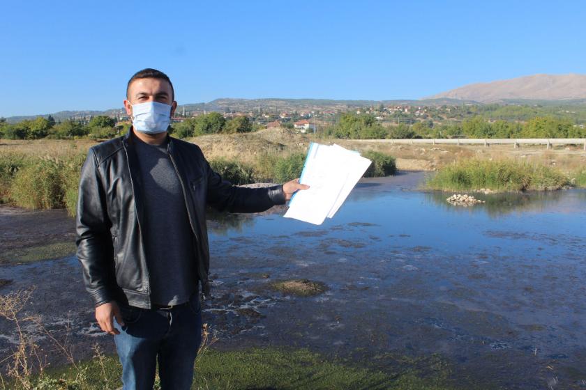 Deniz Arıkan, sol elinde tuttuğu dilekçeleri gösteriyor, arkada kanalizasyon atıklarının olduğu alan