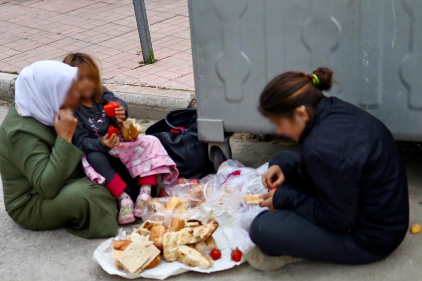 Bir çöp konteynerinin yanında yere oturmuş ekmek ve domates yiyen iki kadın ve bir çocuk.