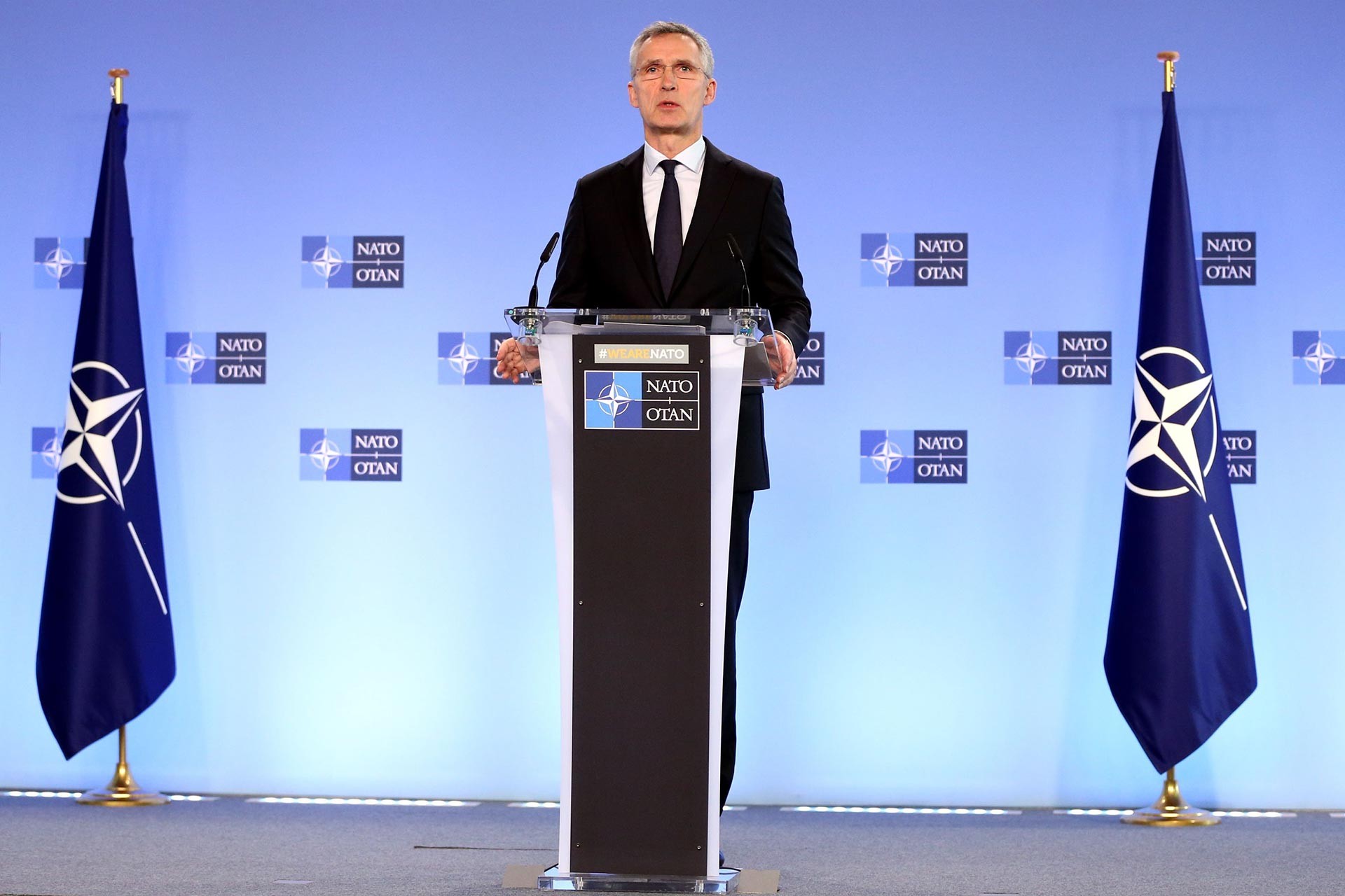 Jens Stoltenberg, iki NATO bayrağı arasında, kürsüden açıklama yapıyor. 