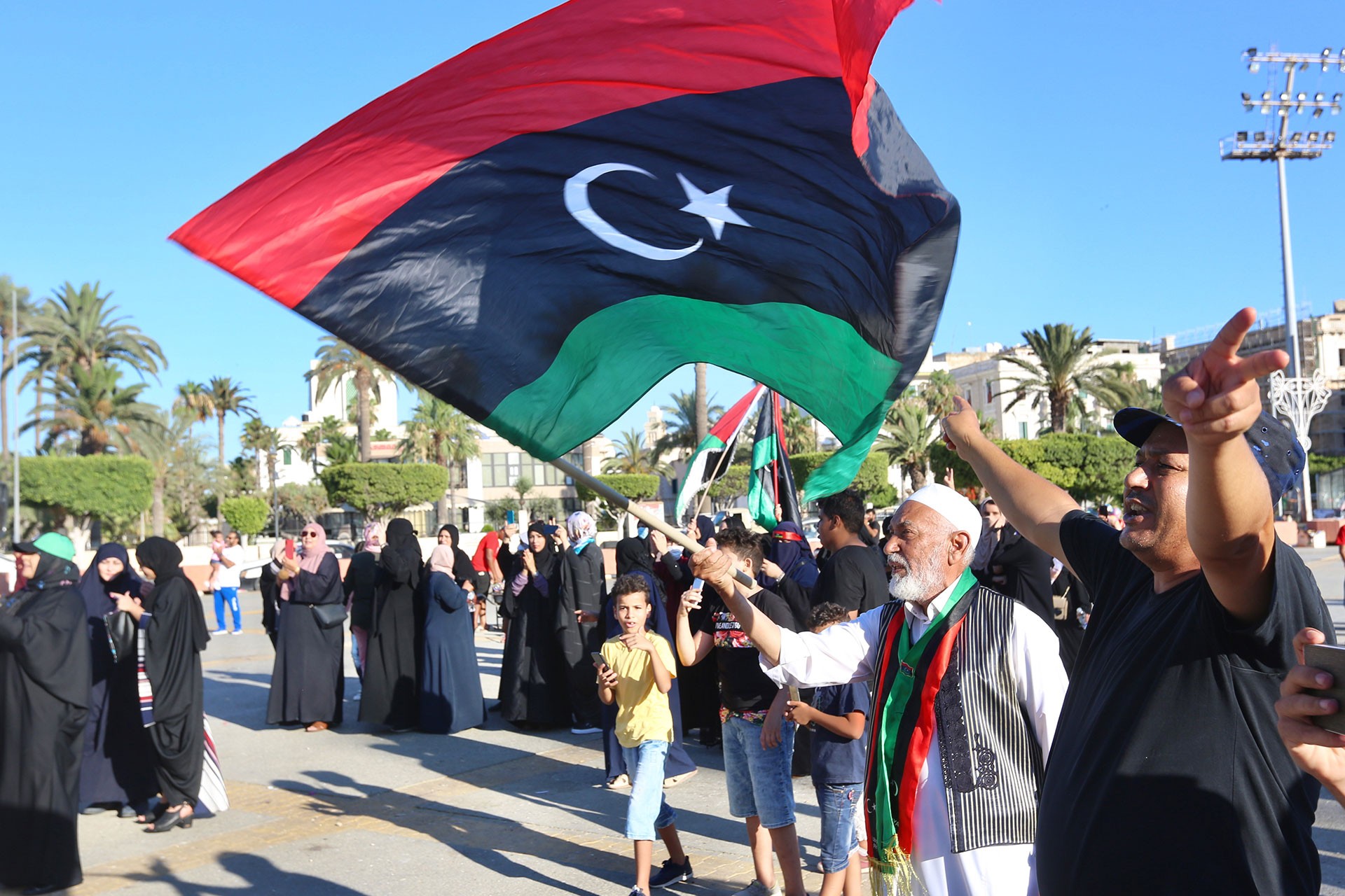 Elindeki Libya bayrağını sallayan yaşlı bir erkek, sağdaki erkek zafer işareti yapıyor